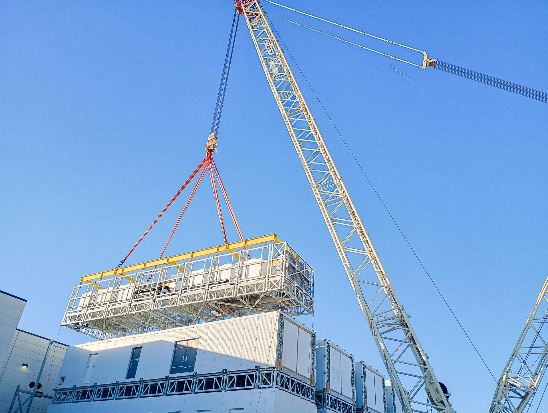 Crane lifting a large, caged structure above a white building against a clear blue sky.