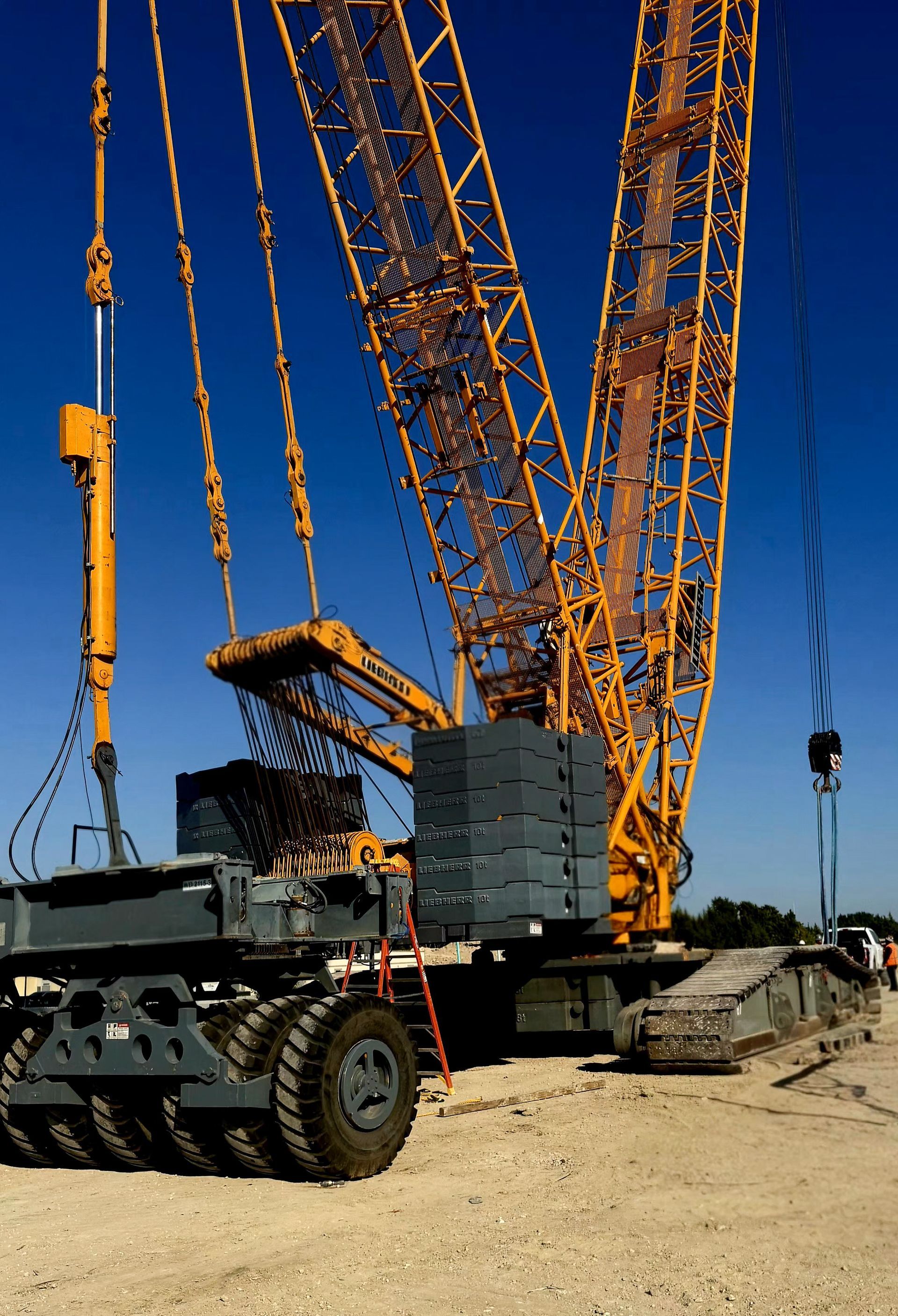 Yellow construction crane against a blue sky, with a tracked base and tires, on a construction site.