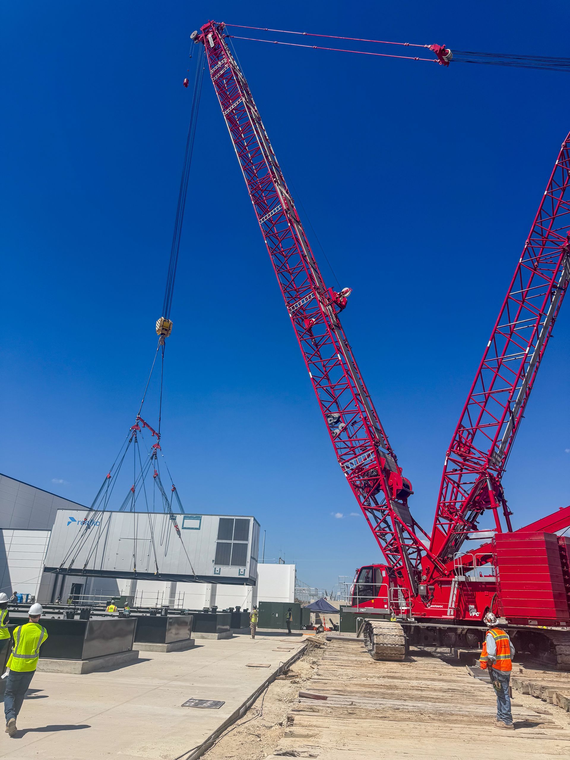 Large red crane lifting a white building module on a construction site. Two workers in safety vests. Bright blue sky.