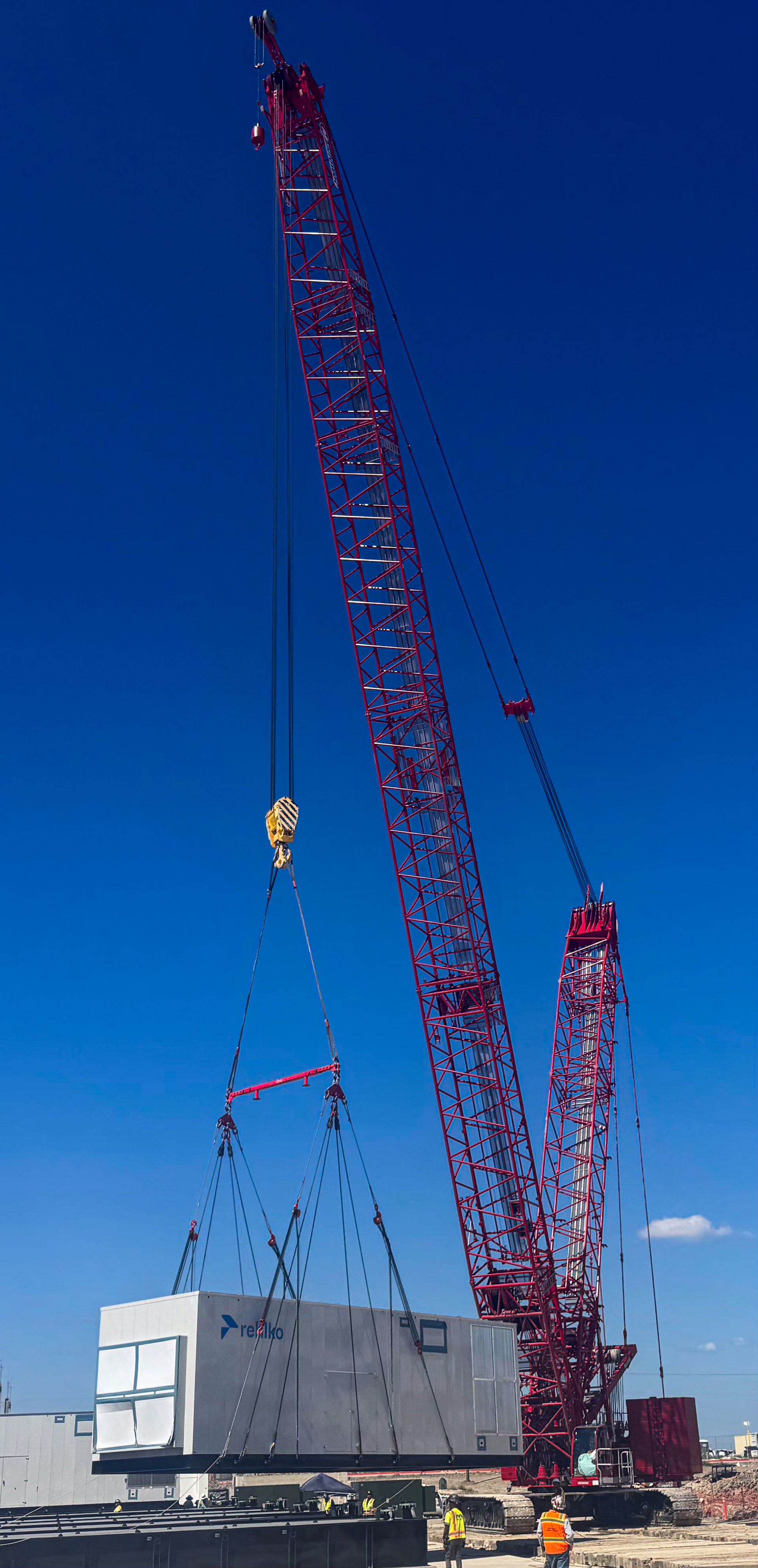 A large red crane lifts a concrete structure against a clear blue sky. Two workers are at the base of the crane.