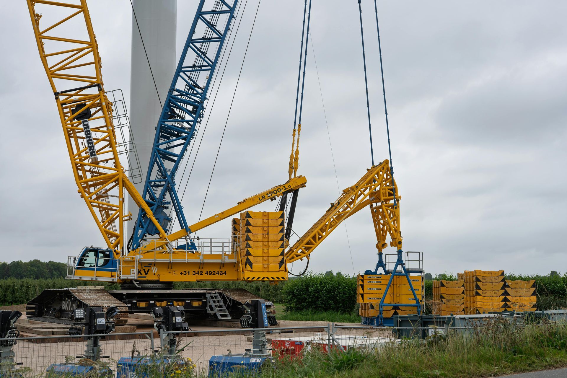 A large yellow and blue crane with a suspended counterweight system, parked at a wind turbine construction site.