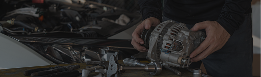 Hands inspecting a car alternator in a dimly lit engine bay. | Dean's Tire & Auto