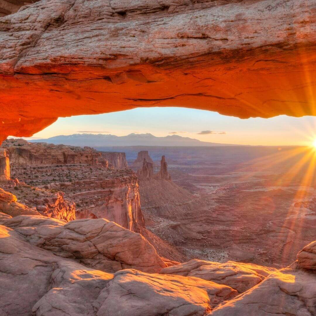 sun rising through an arch in a rocky, mountainous desert