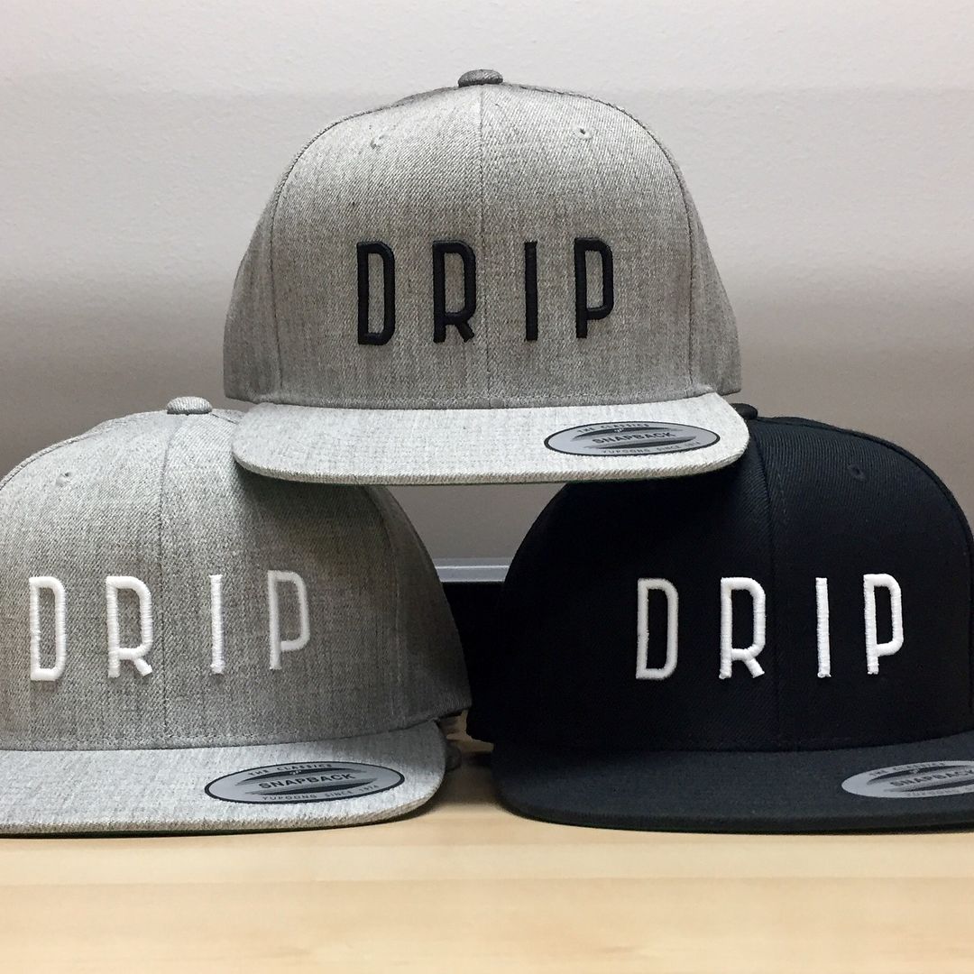 Three caps stacked on a surface, two light grey and one black, all featuring the embroidered word 