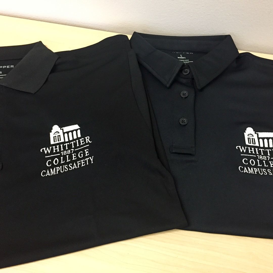 Two black polo shirts featuring the embroidered white Whittier College Campus Safety logo on a light-colored surface.
