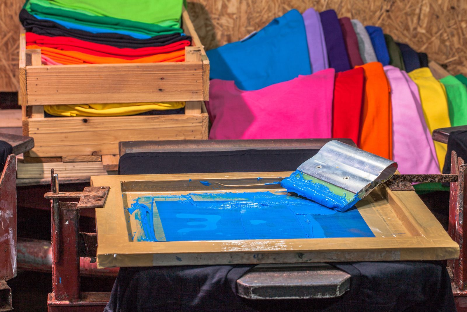 A screen printing frame with blue ink and a squeegee positioned above a black shirt, with stacks of colorful shirts nearby.