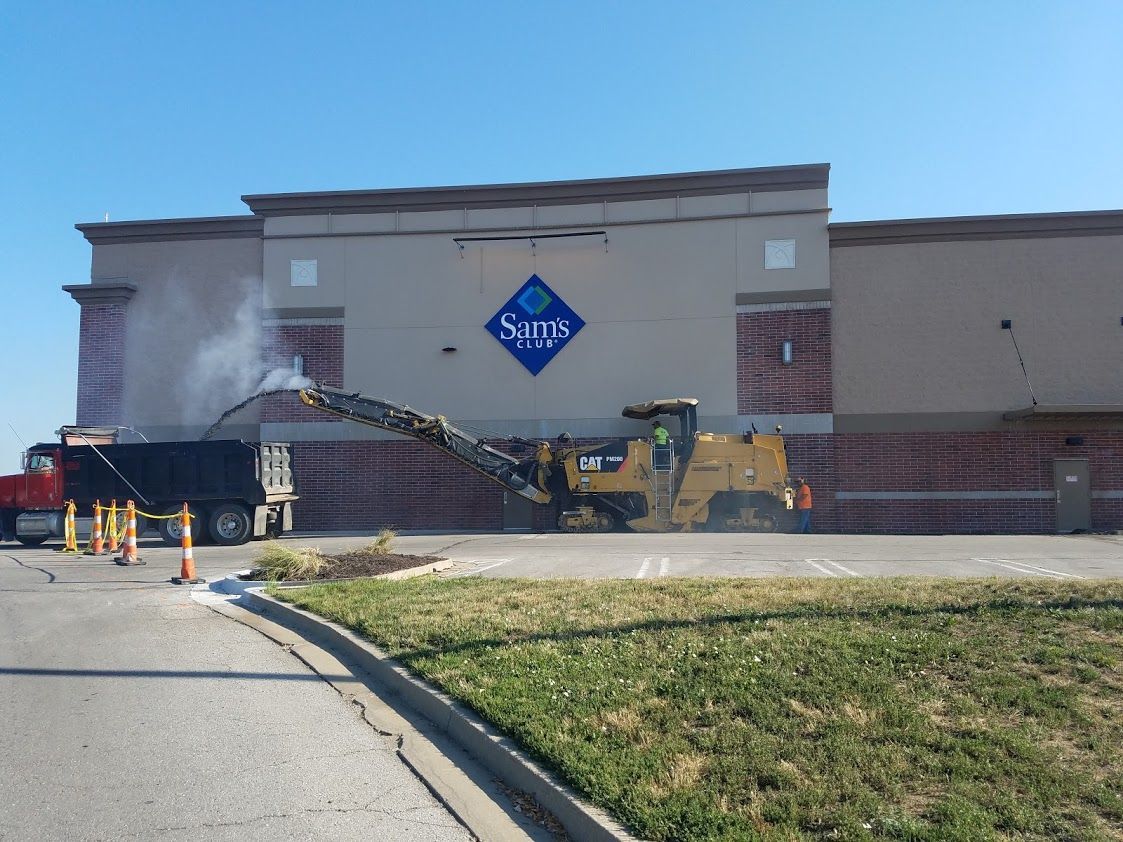A road milling machine is removing pavement in front of a Sam's Club store, loading debris into a truck on a sunny day.
