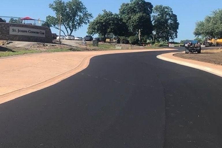 A newly paved asphalt road curves towards a sign for St. Joseph Cemetery. The road is bordered by tan concrete.