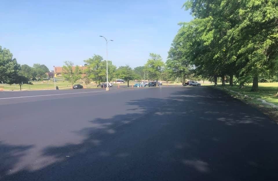 Newly paved asphalt road with cars parked in the distance, bordered by trees and a green grassy area. Blue sky overhead.