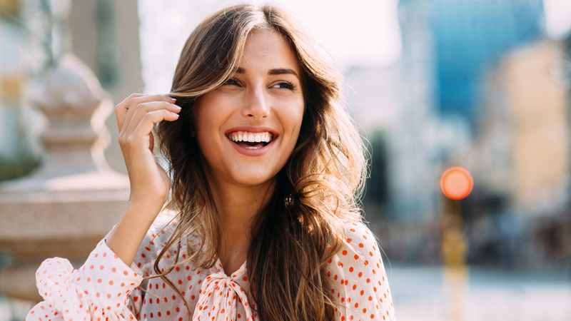 Woman with long brown hair, smiling outdoors, touching hair, wearing polka-dot blouse, sunlit, blurred city background.