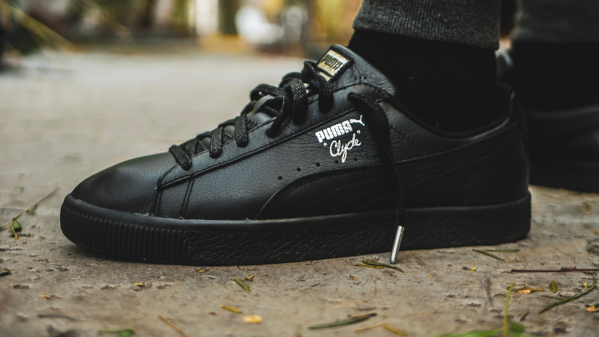 Black Puma Clyde sneaker on a person's foot, standing on a path, near a nail.