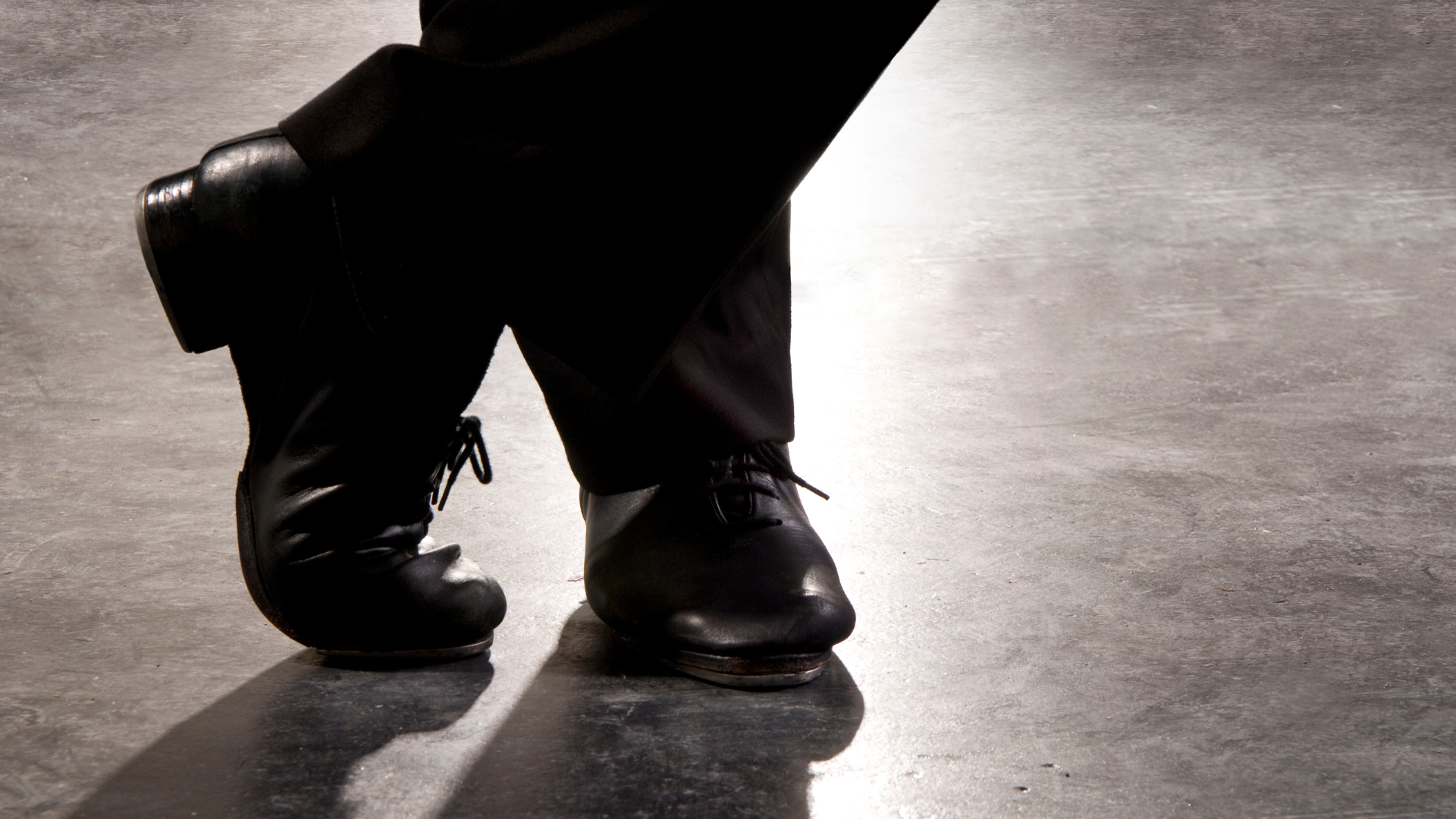 Tap dancer's feet crossed, wearing black tap shoes, on a gray stage.