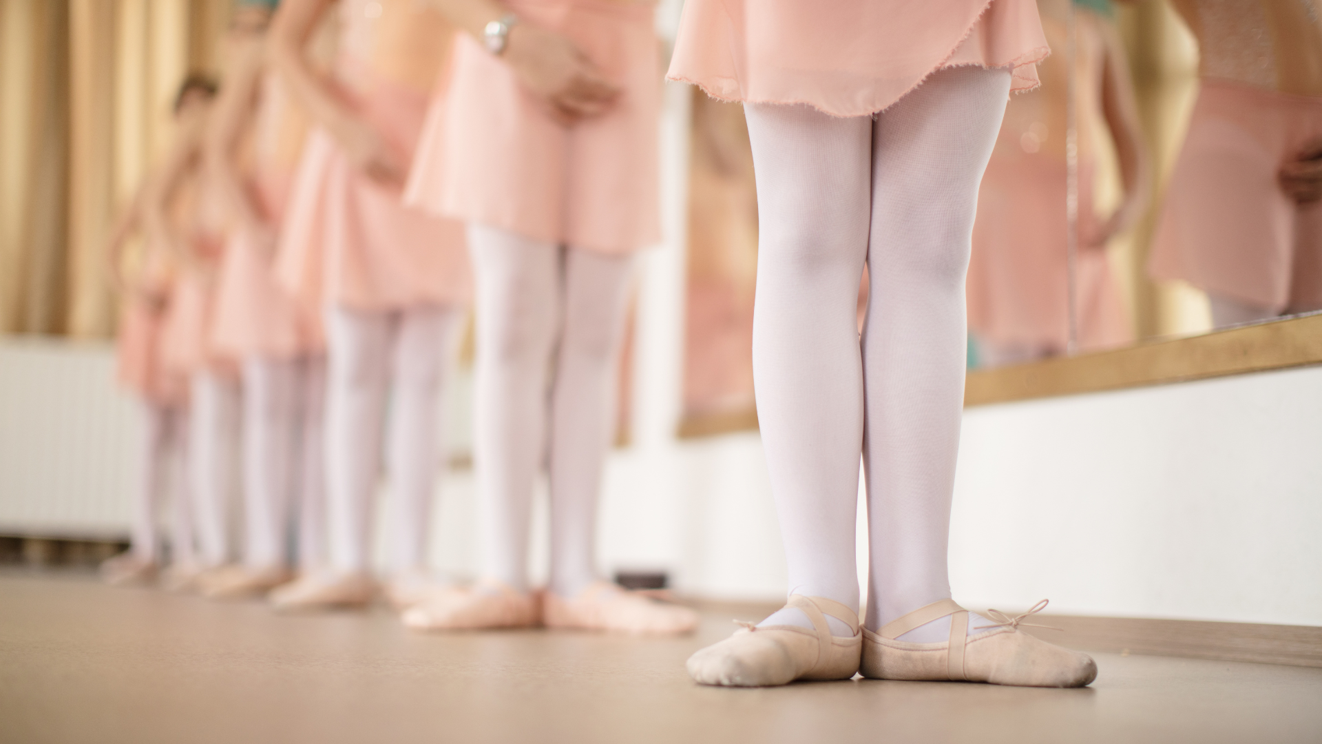 Young ballet dancers in pink outfits and white tights standing in a line near a mirror.