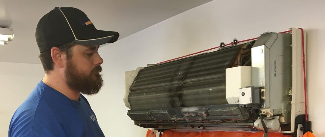 A man with a beard wearing a cap is inspecting an air conditioner unit mounted on a wall.
