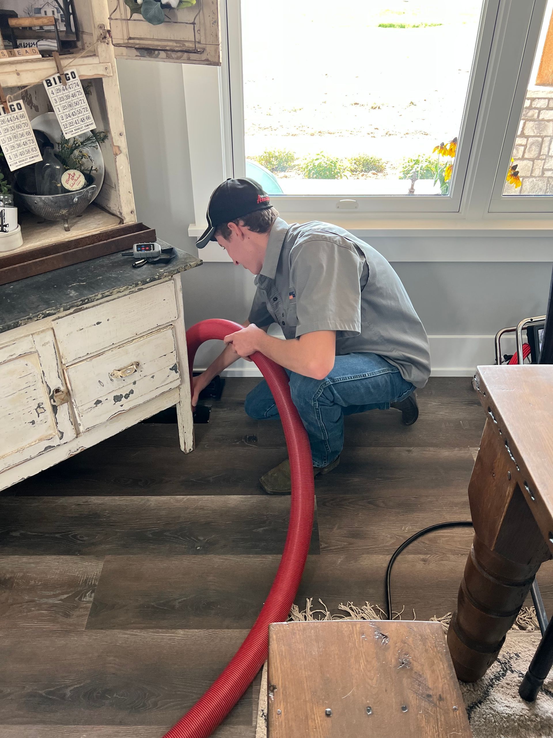 A person in a cap crouches near a red hose cleaning an antique cabinet.