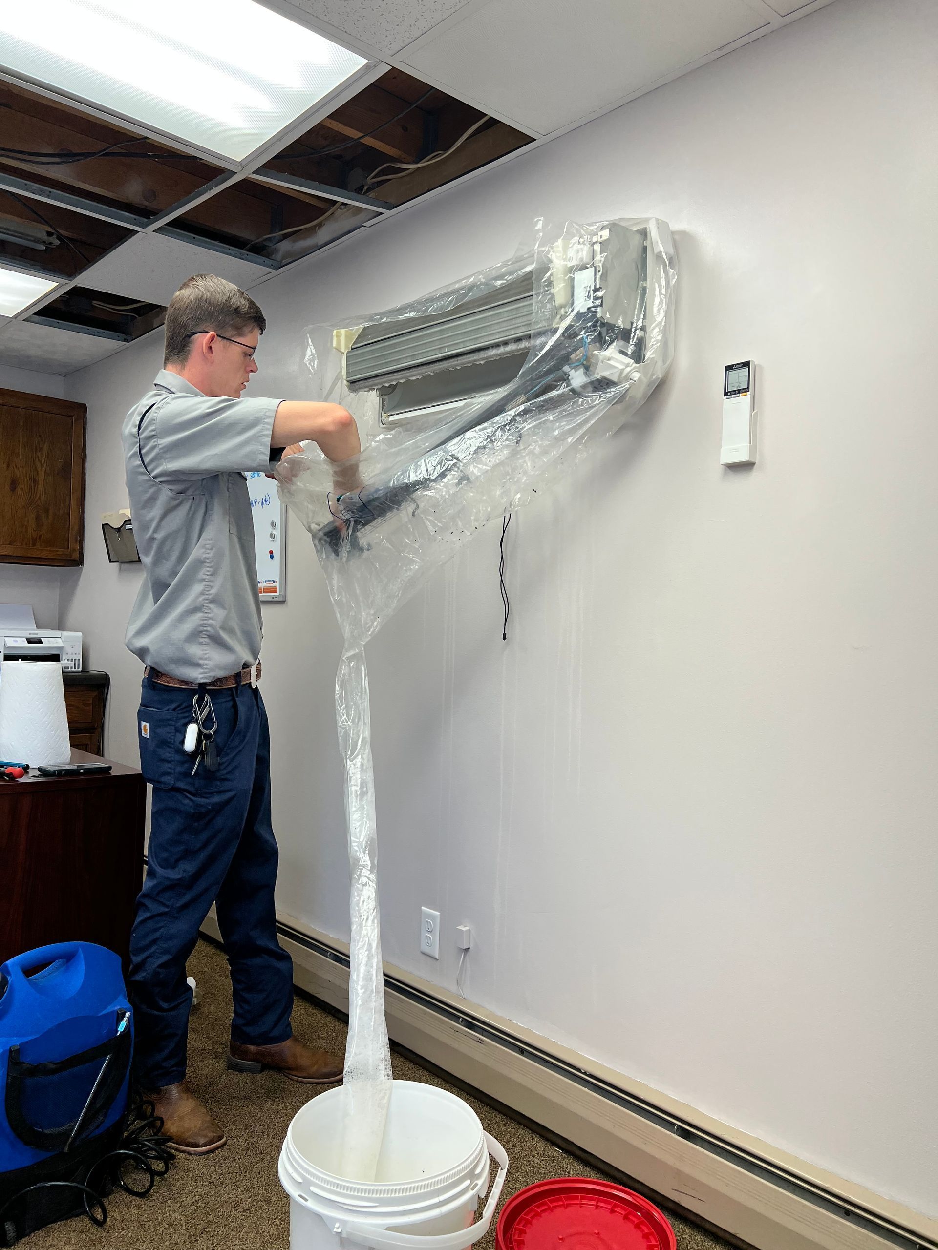 A man installing a ductless AC unit on a wall inside a room, plastic wrap covers the unit and bucket below.