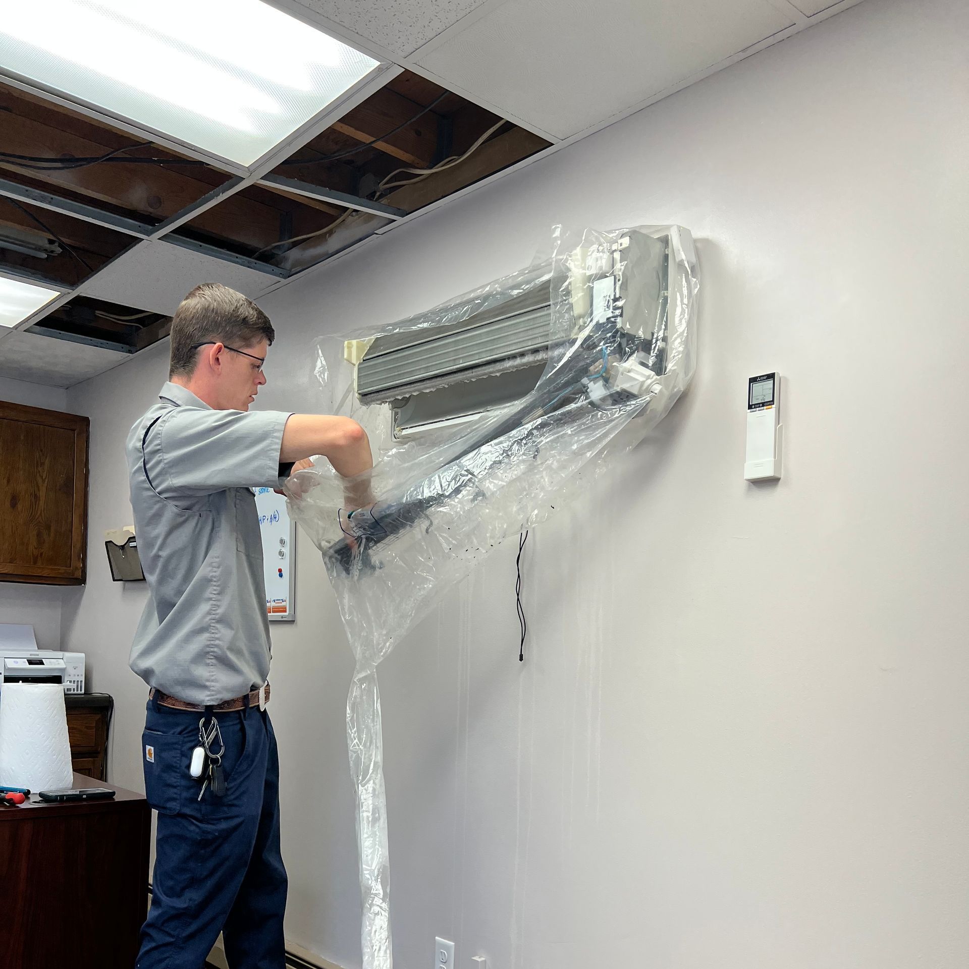 Man in gray shirt installing an air conditioner on a beige office wall.