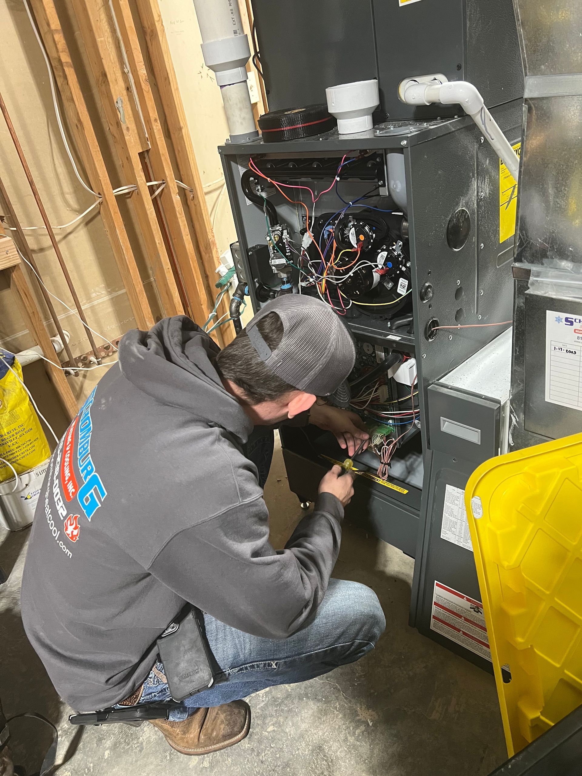 HVAC technician kneeling, working on furnace wiring, in utility room. Wearing gray hoodie and jeans.