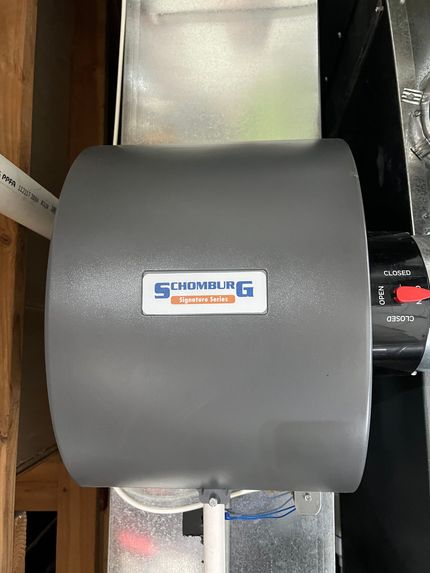 A gray whole-house humidifier attached to ductwork in a wooden-framed utility room.
