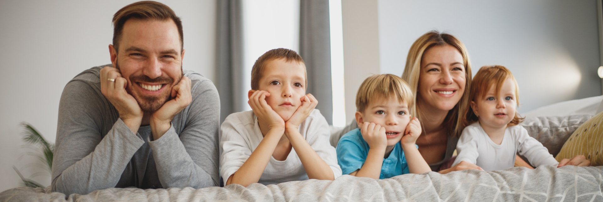 A family of five smiles and rests their chins on their hands, lying on a sofa.