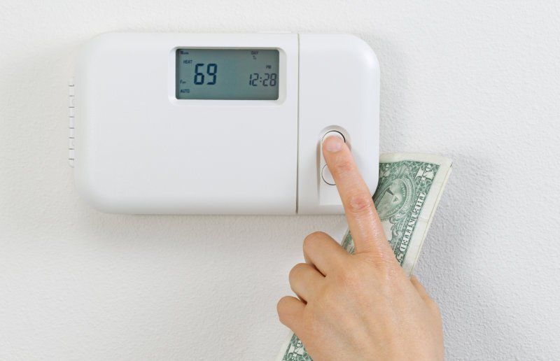 Hand pressing thermostat button with a dollar bill, showing 69 degrees.