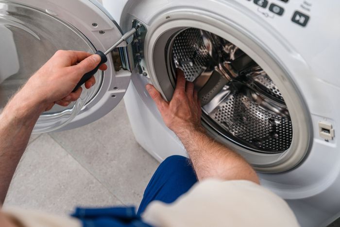 A technician in a uniform repairing the metal door hinge of a white front-loading washing machine with a screwdriver.