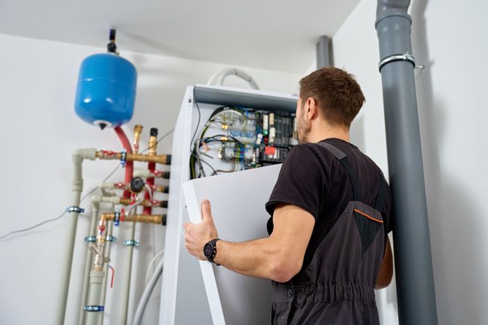 A technician in work coveralls removes the front panel of a wall-mounted heating boiler in a white utility room.