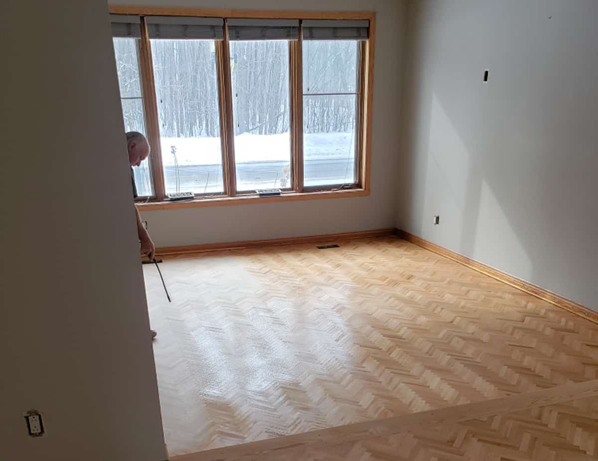 Man refinishing hardwood floor in a room with large windows, snow visible outside.