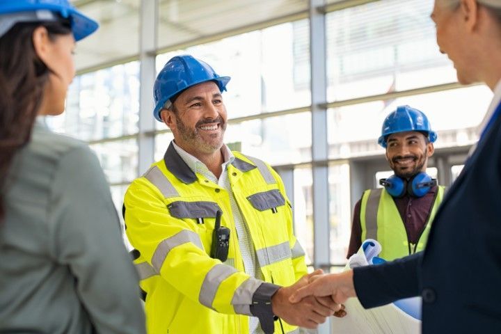 Construction workers in hard hats shaking hands on a job site, one wearing a bright yellow jacket.