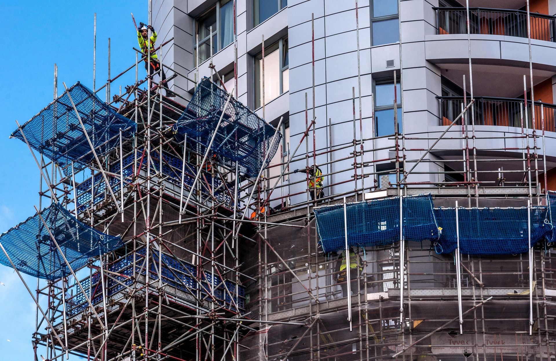 Scaffolding surrounds a curved building under construction, with blue safety netting against a clear sky.