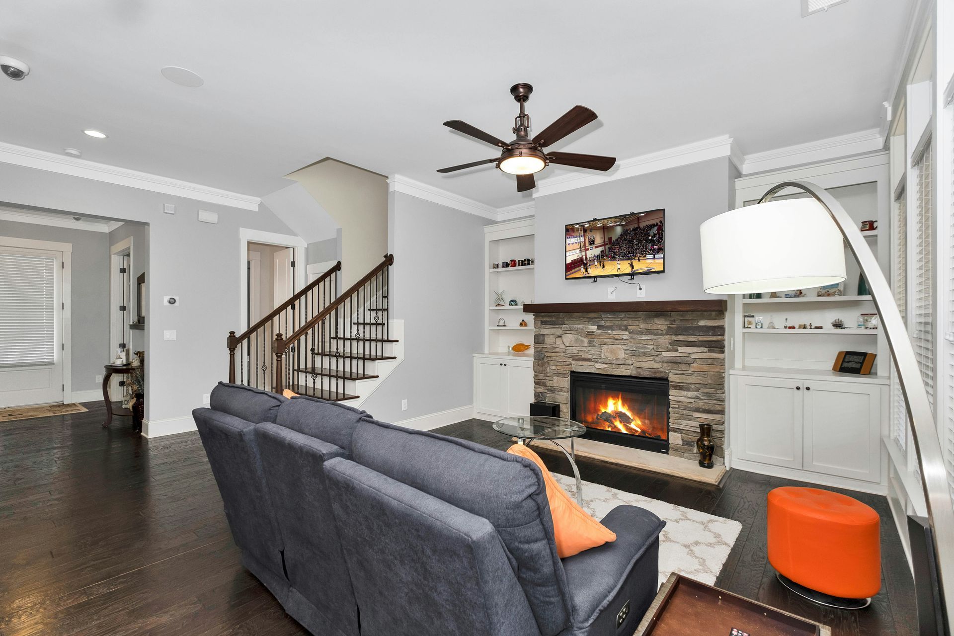 Modern living room with gray sofa, fireplace, ceiling fan, staircase, and built-in shelves