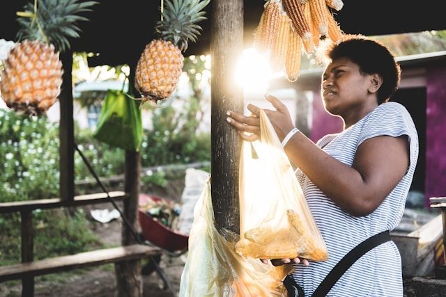 A woman is standing in front of a pineapple stand holding a bag of corn.