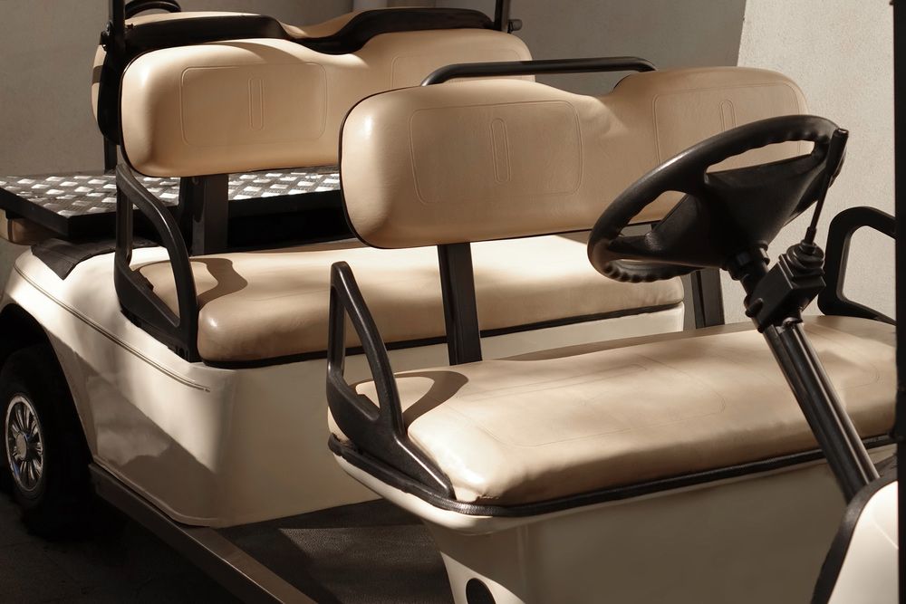 Close-up of a beige golf cart with two rows of seats, steering wheel, and black handrails