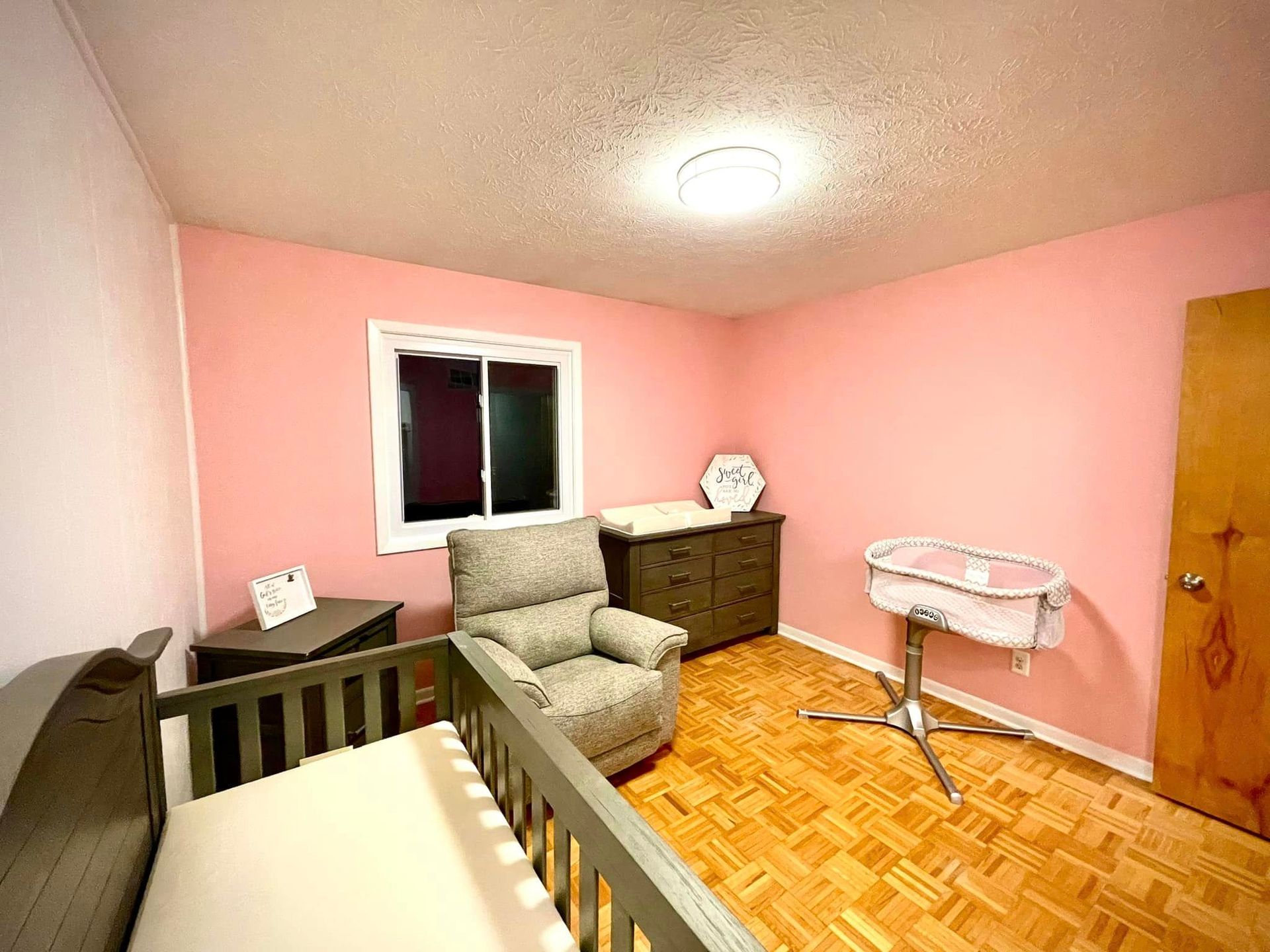 A baby room with pink walls , a crib , a chair , a dresser and a window.