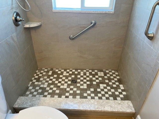 Shower with gray tiled walls and a checkerboard tile floor, featuring a grab bar and a step up.