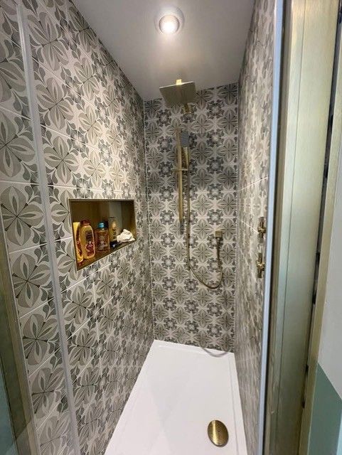 Small shower with patterned tile walls, gold fixtures, and a built-in shelf for toiletries.