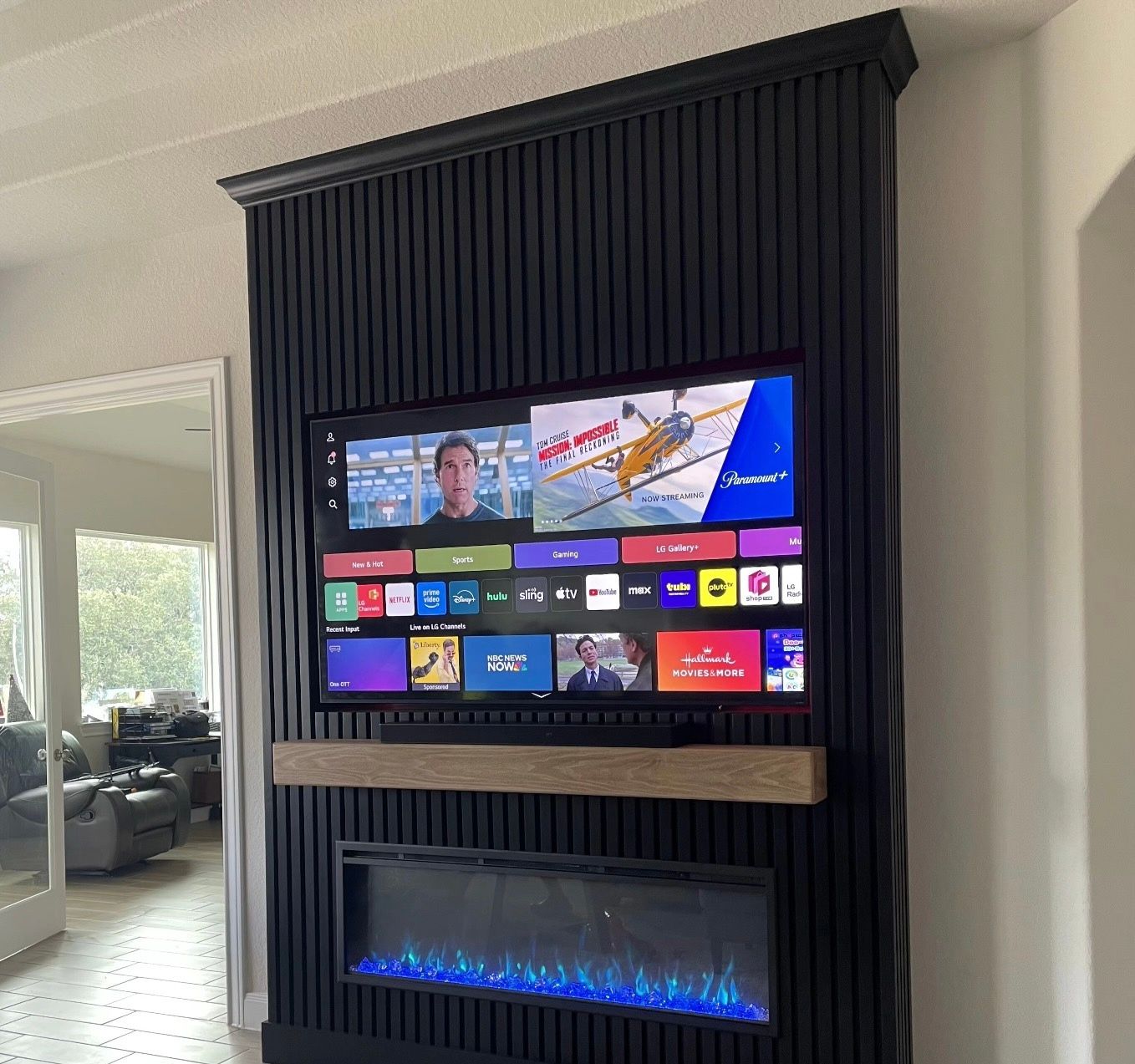 Black-paneled wall with a TV and fireplace. The TV displays a streaming service. A wooden shelf sits above the fireplace.