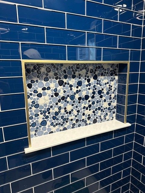 Blue tiled shower niche with pebble tile back and gold trim. White shelf.