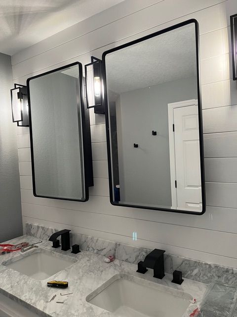 Two framed mirrors above double sinks in a bathroom. Black faucets, white countertop, white shiplap walls.