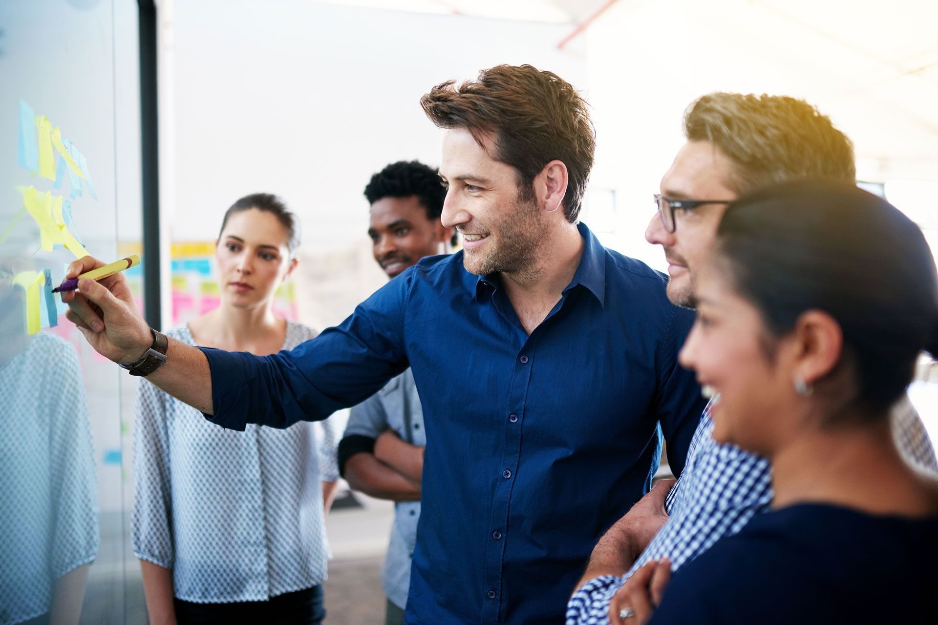 Group of people looking at a whiteboard with a man drawing on it; sunny office.