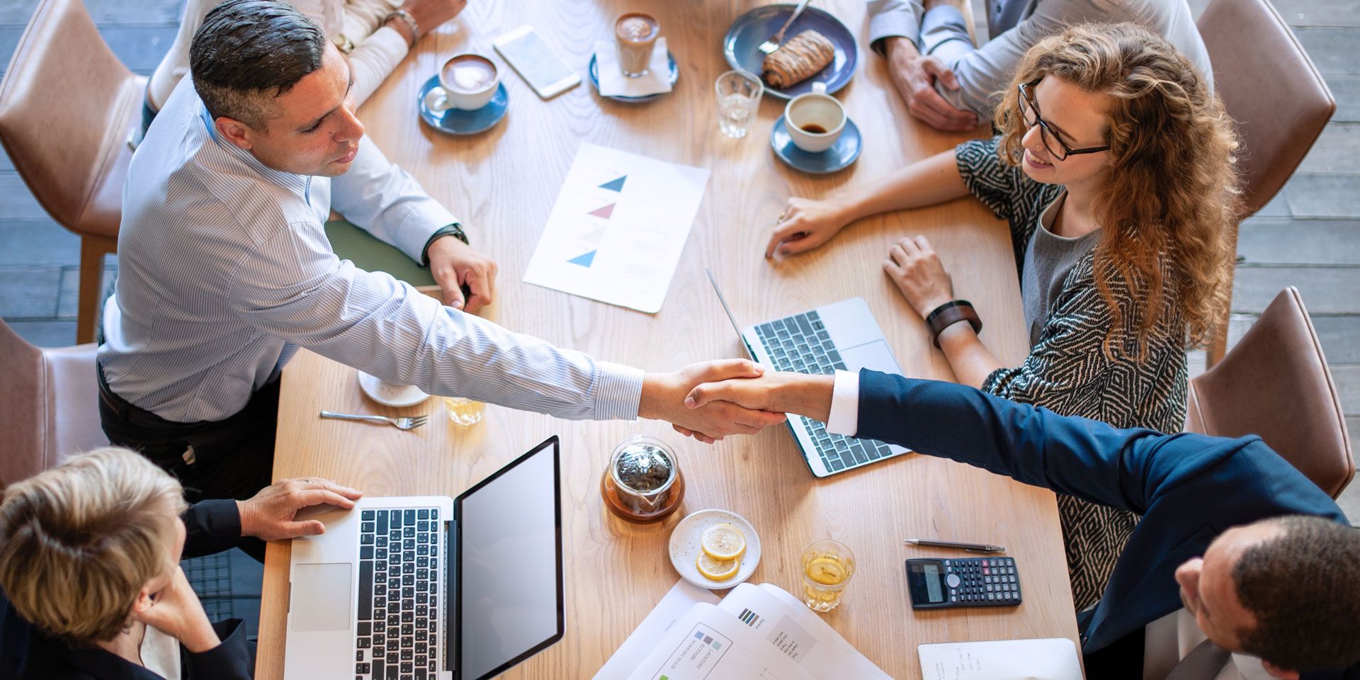 Business meeting around a table. Two men shaking hands, others watching, with laptops and documents.