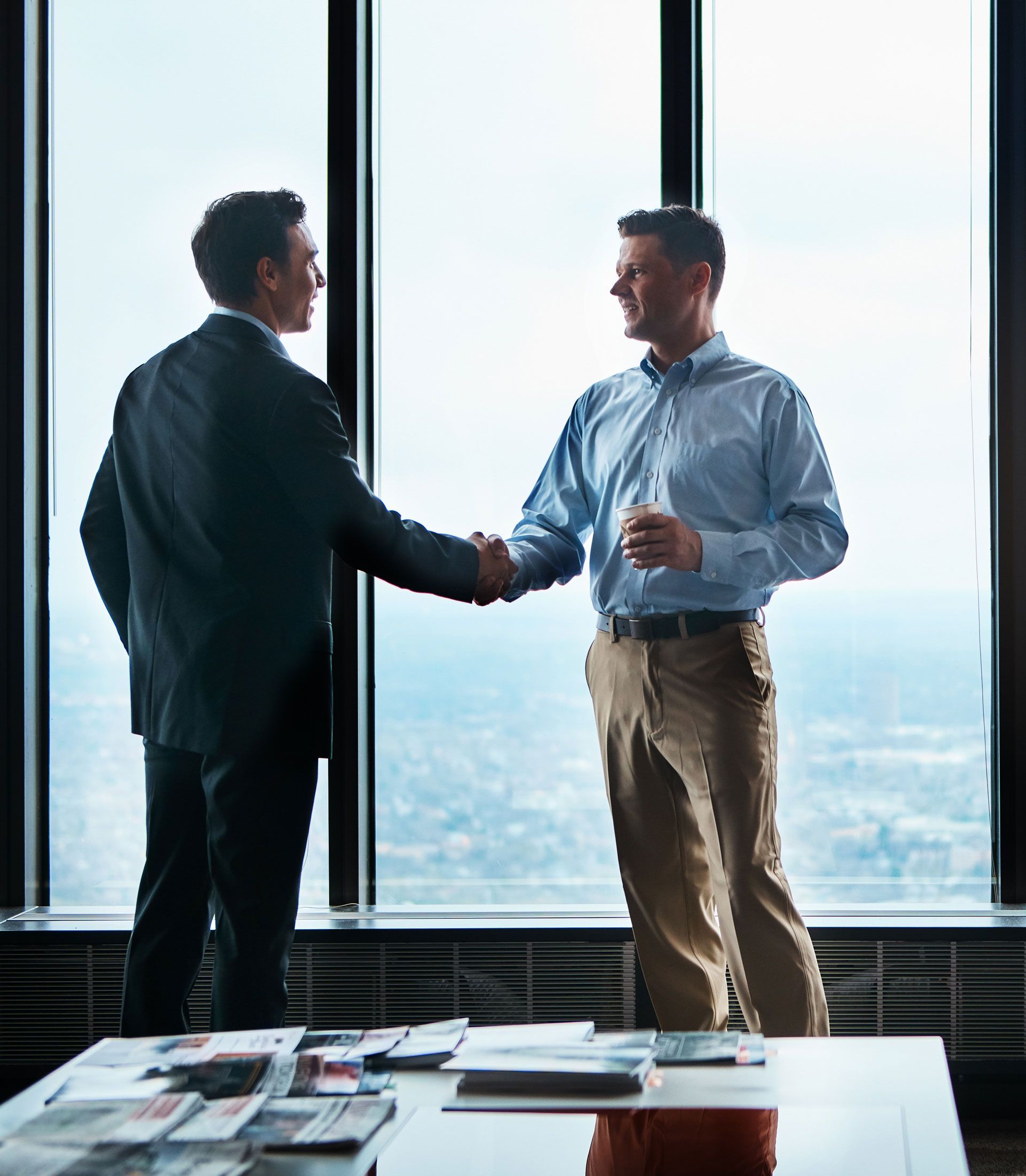 Two men shake hands in front of a window overlooking a cityscape. One in a suit, the other in a button-down shirt.