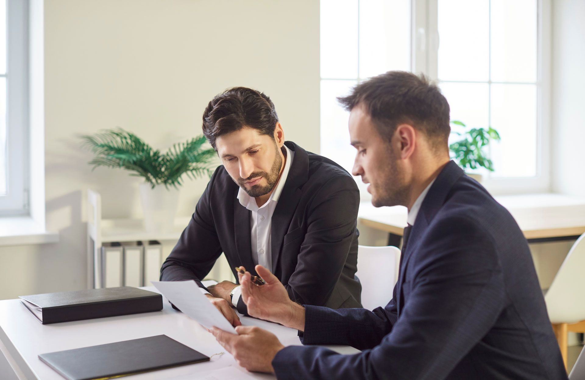 Two men in suits reviewing documents at a table; one points, the other looks intently.