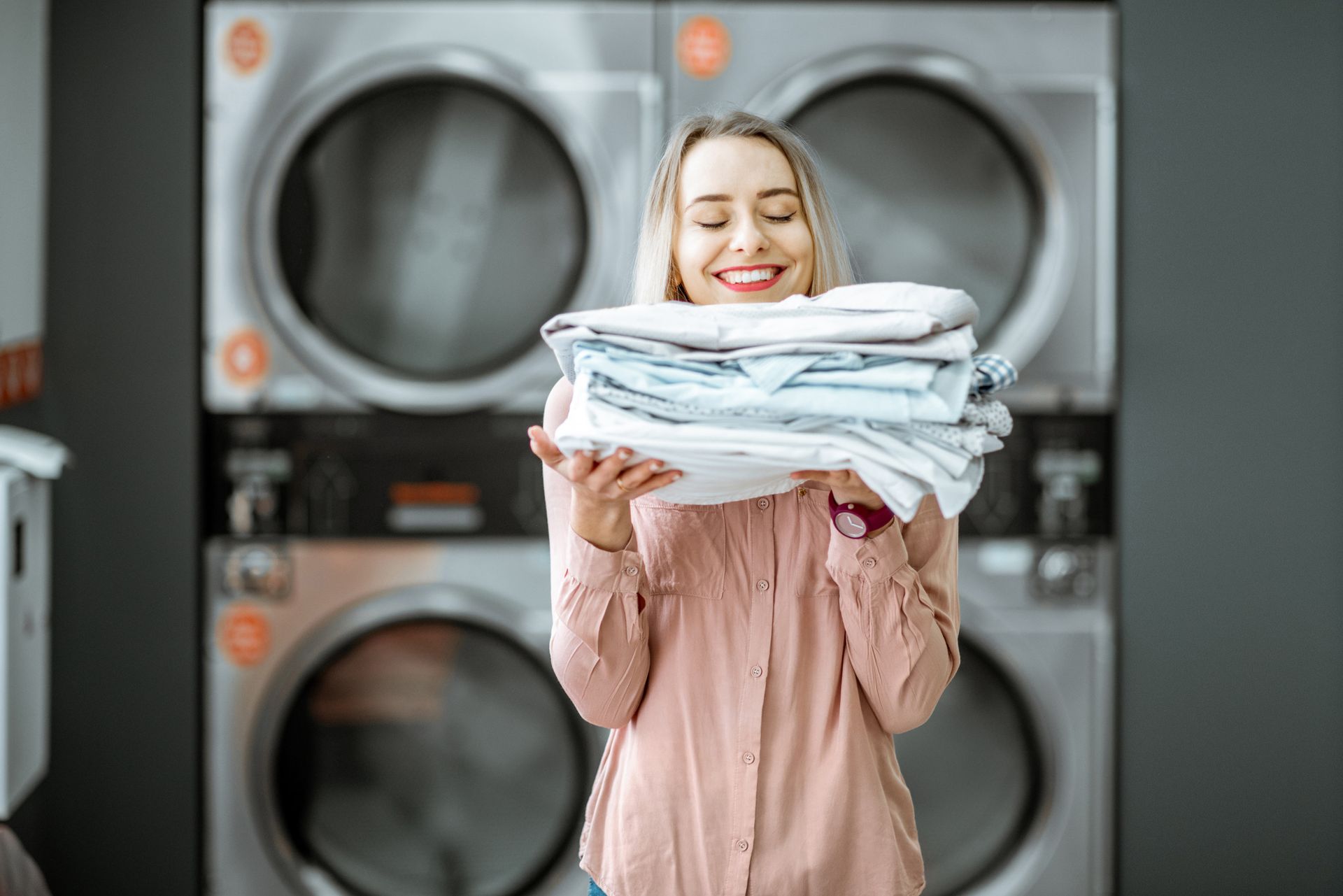 Woman smiling, holding a stack of clean laundry in front of washing machines.
