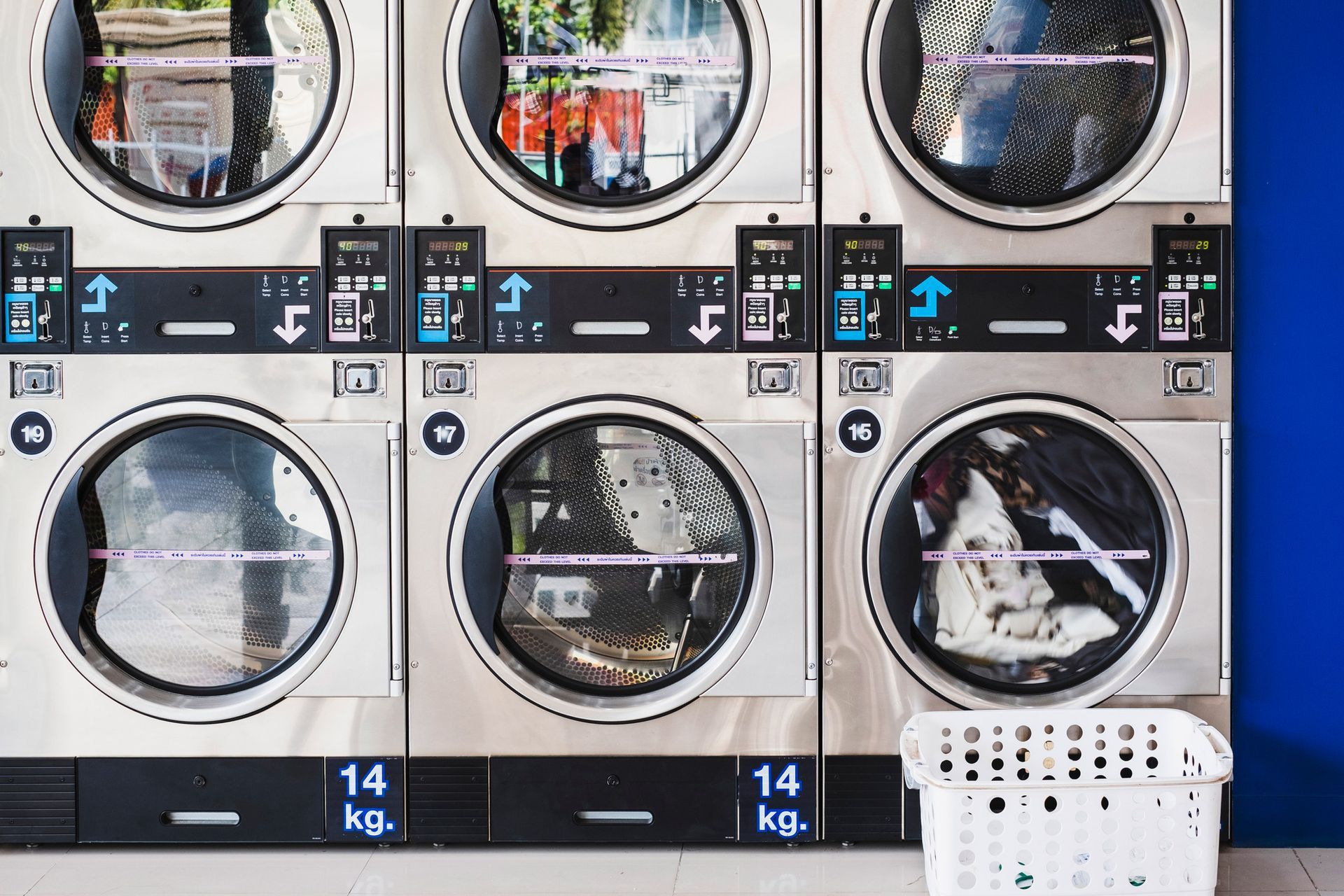 Row of silver washing machines at a laundromat; one machine contains clothes, white laundry basket at right.