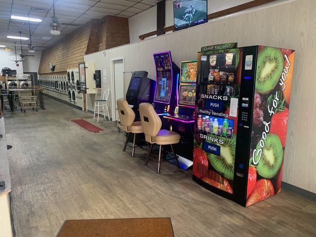 A laundromat with a game area. A vending machine, arcade games, and seating are in the foreground.