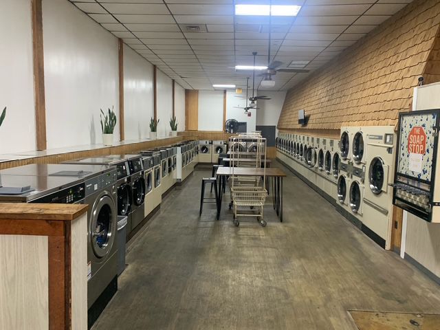 Interior view of a laundromat with rows of washing machines. Empty, with tables and a promotional sign.