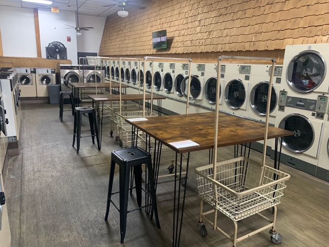 Interior of a laundromat with rows of washing machines, tables, stools, and rolling laundry baskets.