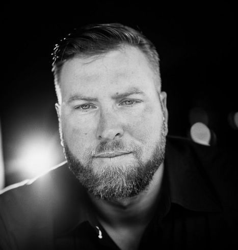 Man with beard looking at the camera in black and white, outdoors, with a soft focus background.