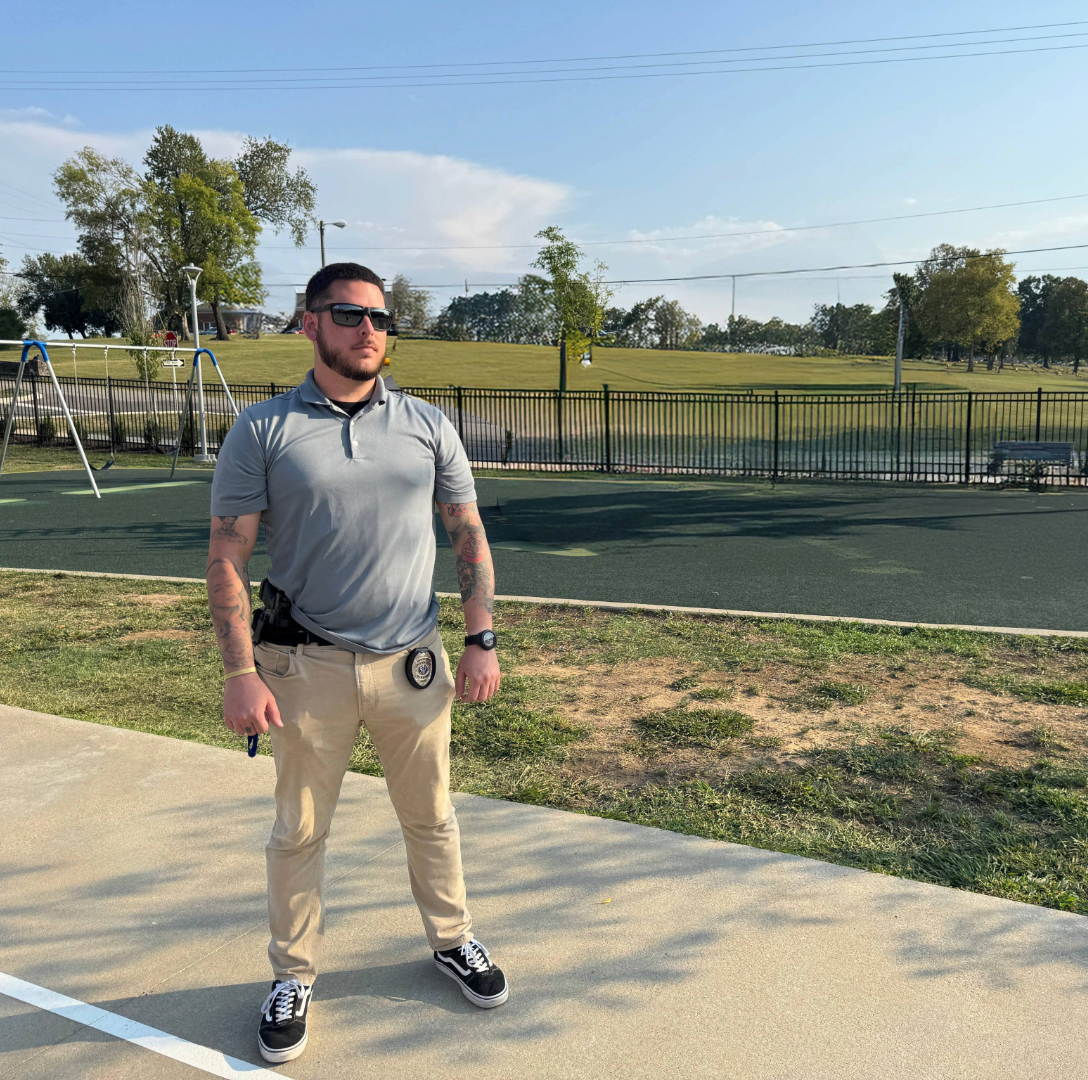 Man with tattoos wearing sunglasses, gray polo shirt, and khaki pants stands outside.